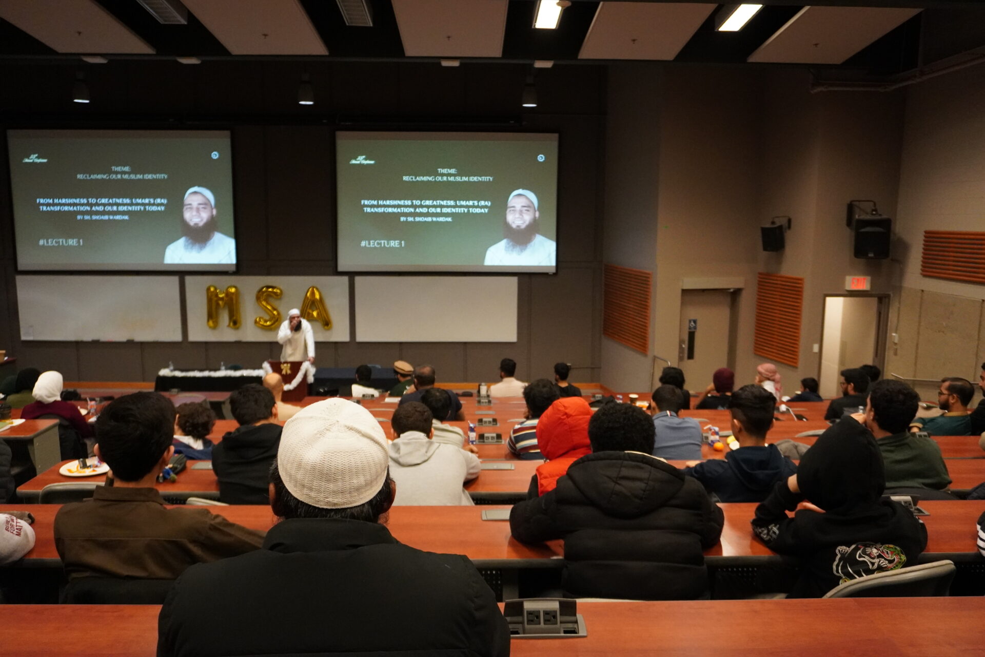 Speaker Sheikh Shoaib Wardak addresses a full lecture hall at the University of Manitoba during the UMMSA 35th Annual Conference, with students and community members seated in tiered rows. Two screens behind the speaker display the conference theme, “Reclaiming Our Muslim Identity."