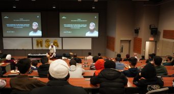 Speaker Sheikh Shoaib Wardak addresses a full lecture hall at the University of Manitoba during the UMMSA 35th Annual Conference, with students and community members seated in tiered rows. Two screens behind the speaker display the conference theme, “Reclaiming Our Muslim Identity."